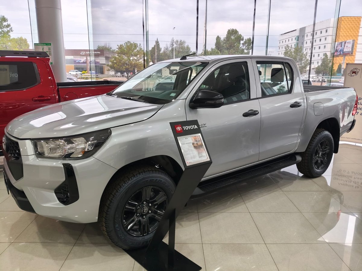 White Toyota pickup truck on display in a dealership showroom with large windows and red vehicle visible in background