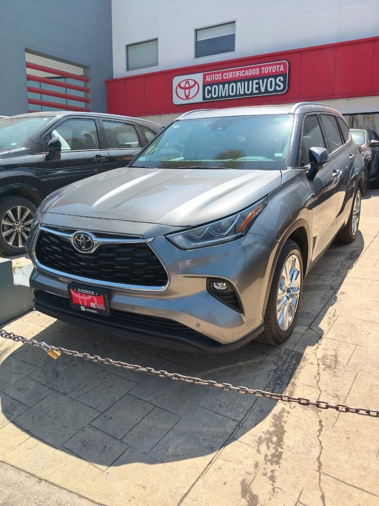 Silver Toyota Highlander SUV displayed at a Toyota dealership with red signage, other vehicles visible in background