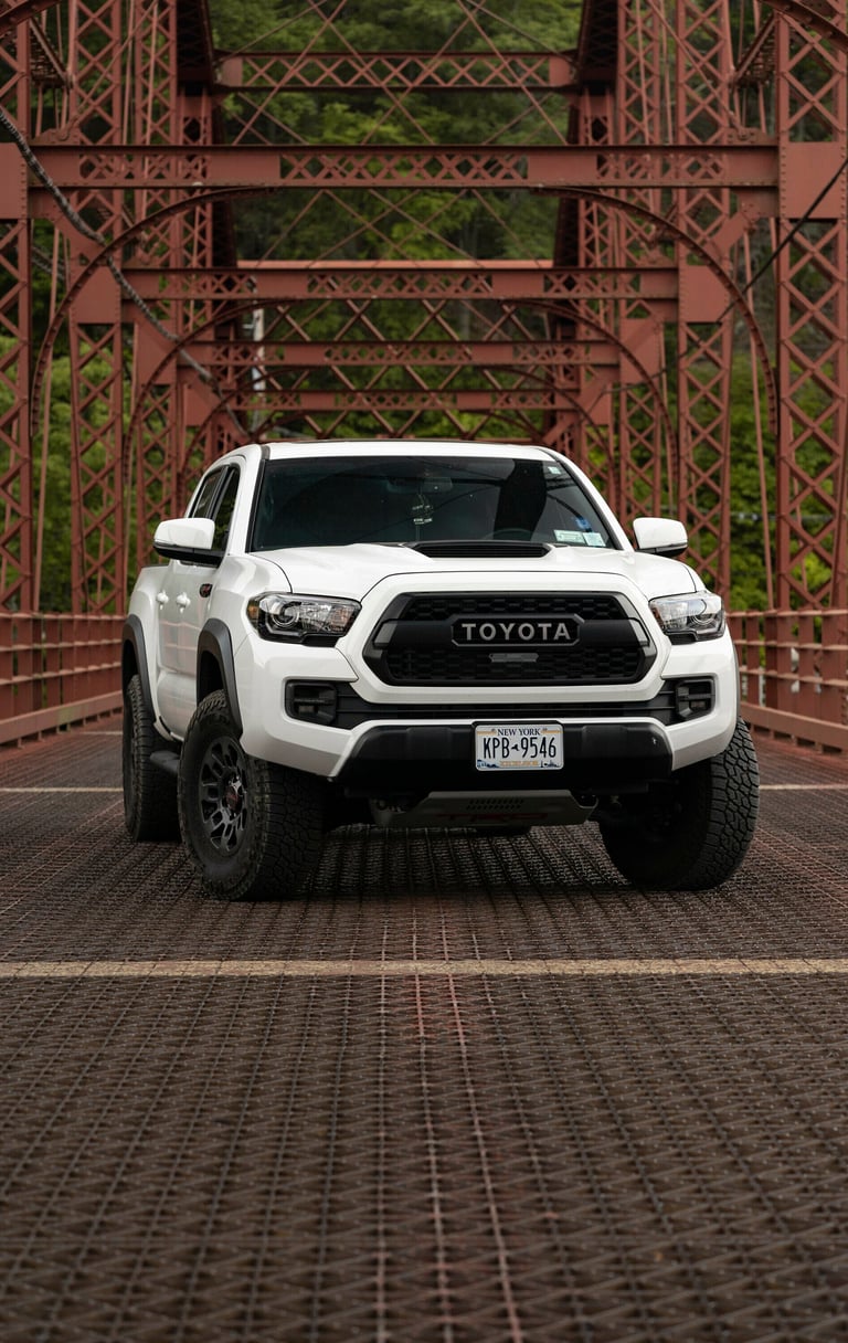 White Toyota Tacoma truck parked on brick bridge under red steel trestle structure with green foliage