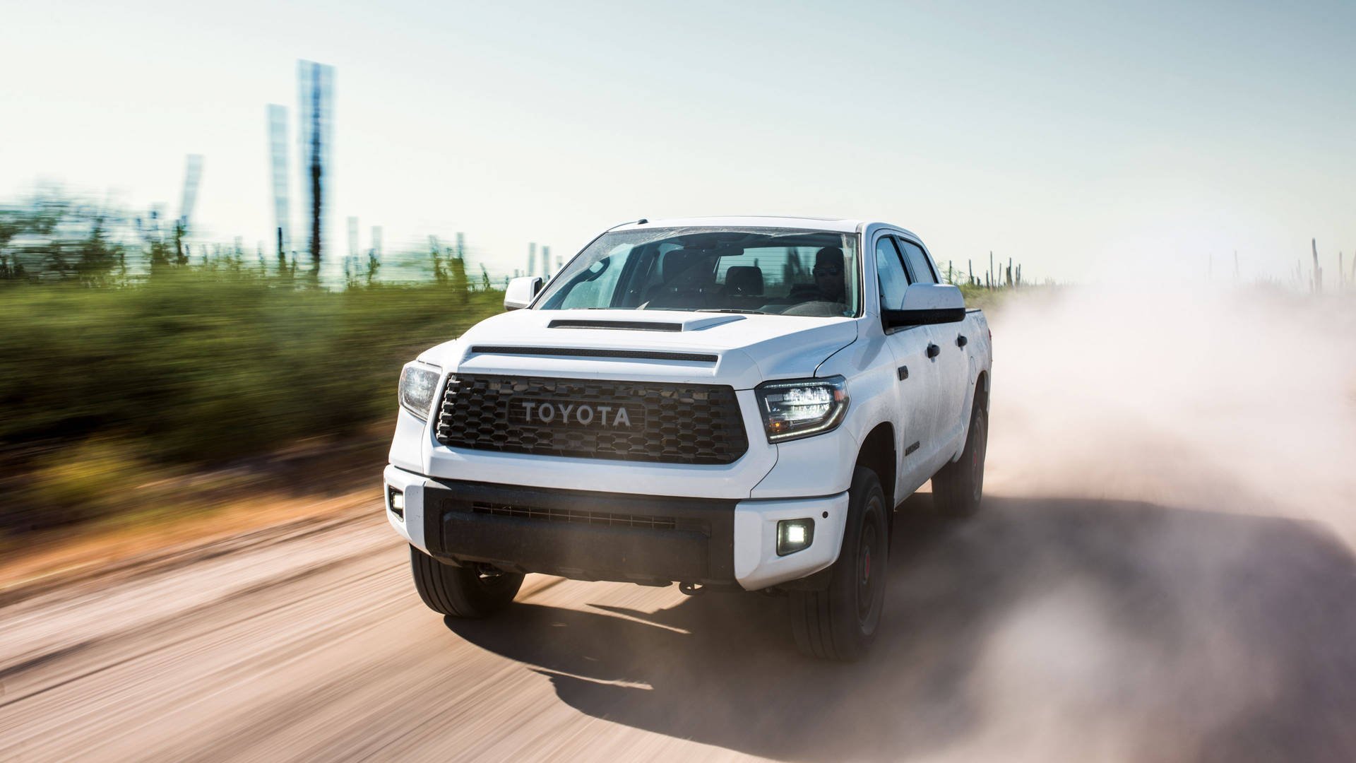 White Toyota pickup truck driving on a dusty dirt road with desert landscape and mountains in the background