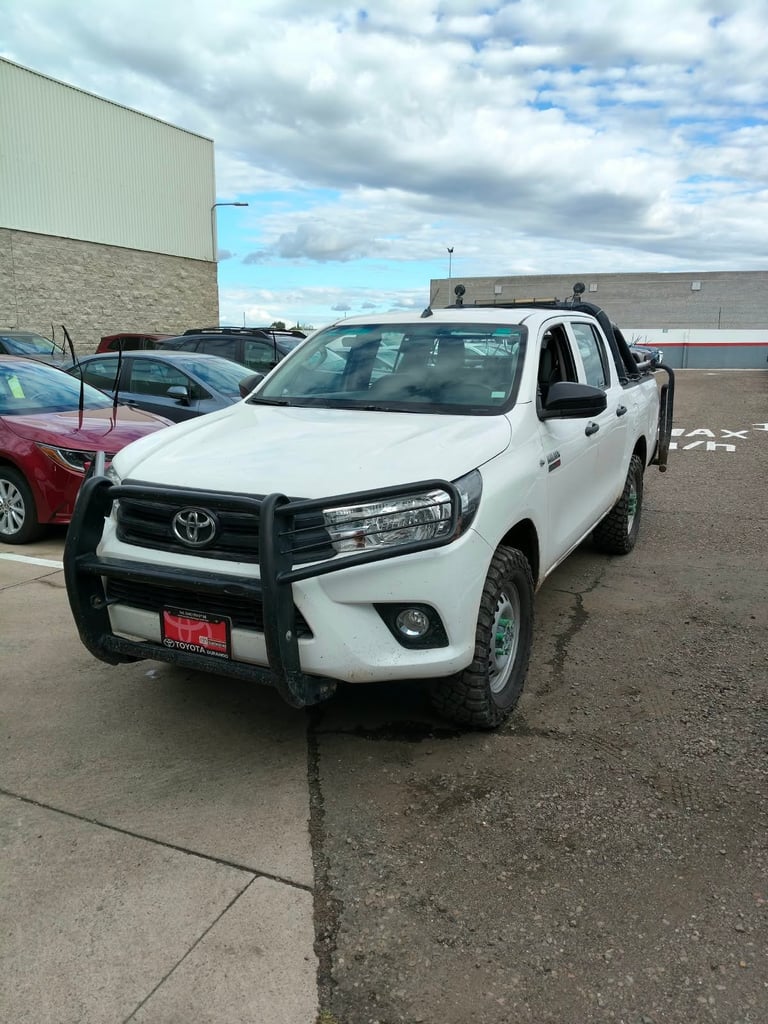 White Toyota pickup truck with black grille guard parked in a lot next to other vehicles and industrial buildings