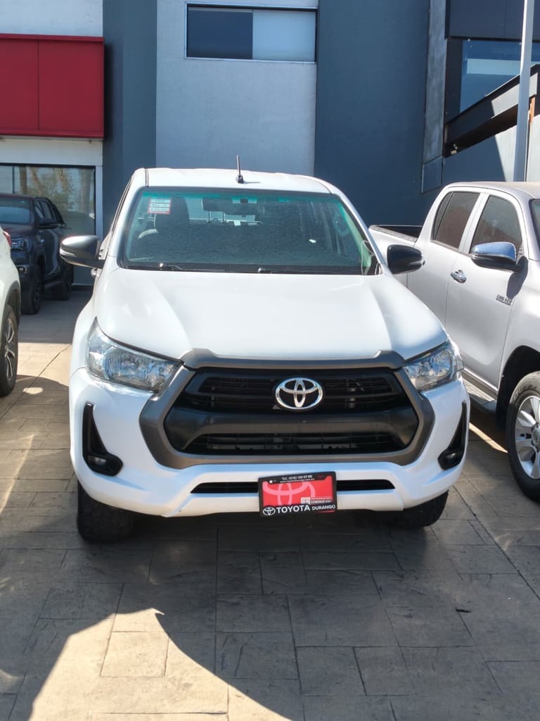 White Toyota pickup truck parked in dealership lot, front view with black grille and red license plate frame